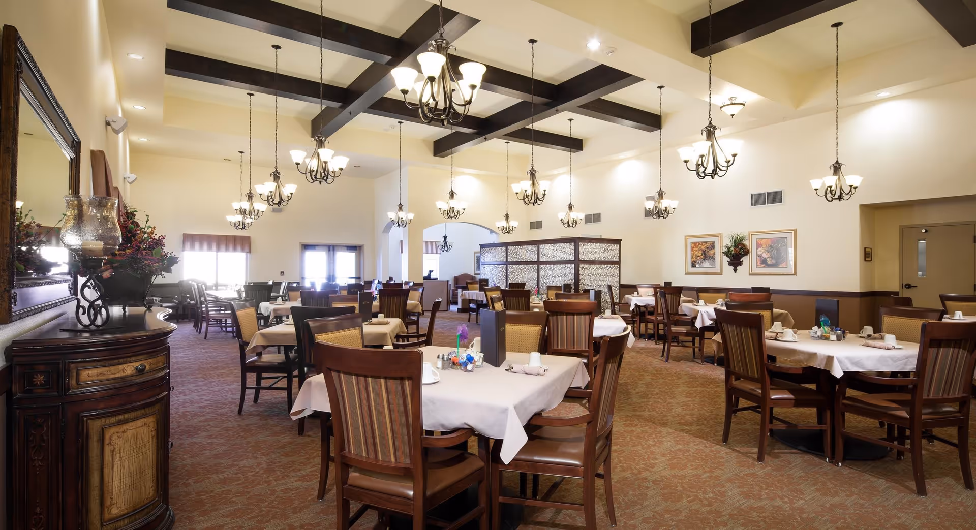 A spacious dining room in a senior living facility with multiple tables covered in white tablecloths, each set with cups, napkins, and condiments. The room features high ceilings with dark wooden beams and multiple hanging chandeliers providing warm lighting. There are framed pictures and decorative plants on the walls, and a large mirror above a wooden sideboard with floral arrangements on the left side.