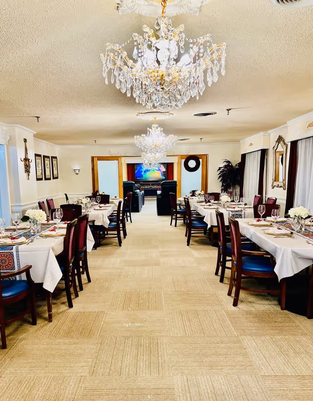 A bright and elegant dining room with tables set for a meal, featuring white tablecloths, floral centerpieces, and wine glasses. The room has crystal chandeliers hanging from the ceiling, framed artwork on the walls, and large windows with curtains. At the far end, there is a seating area with recliners facing a television.