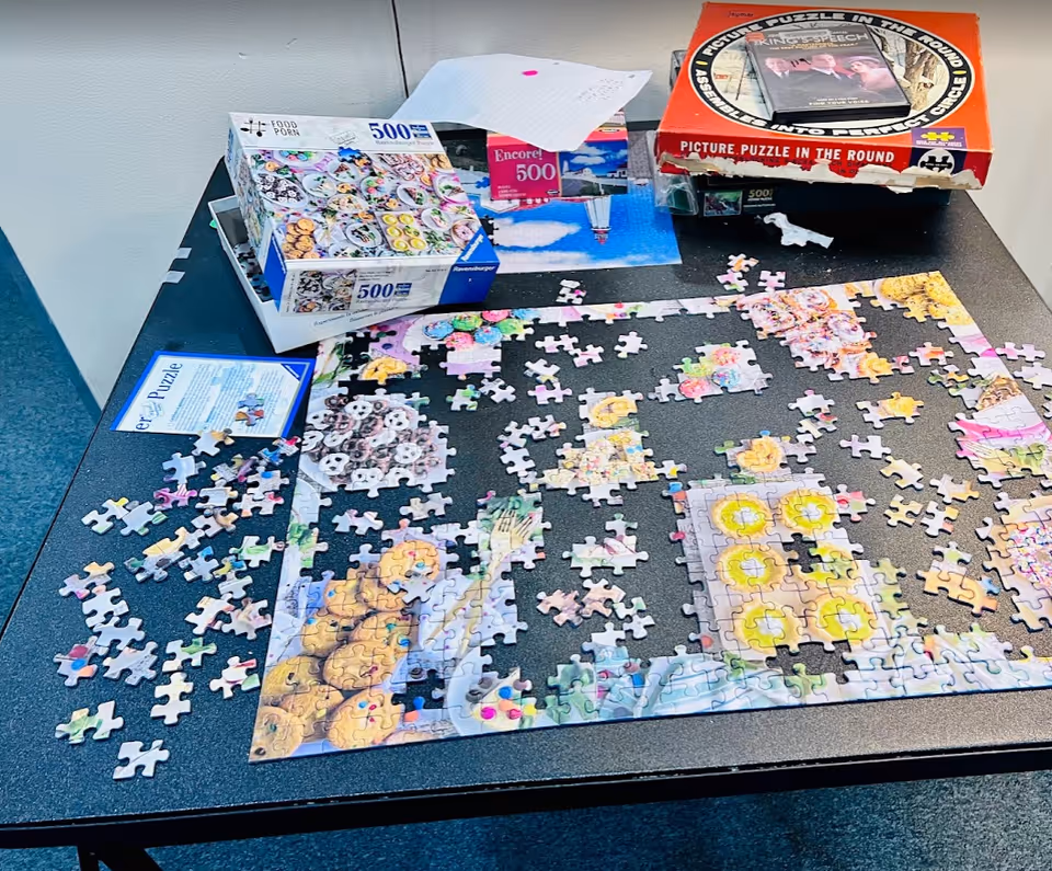 A partially completed jigsaw puzzle on a black table with scattered puzzle pieces around it. Several puzzle boxes and a DVD case are also on the table. The puzzle appears to depict colorful images including food items.