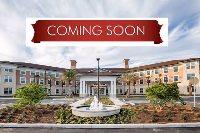 Front entrance of a multi-story senior living building with a fountain and landscaped driveway beneath a 'COMING SOON' banner.