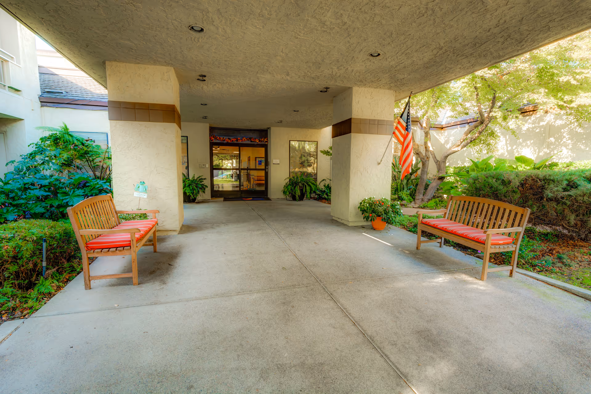 Covered entrance/portico with two wooden benches, potted plants, and an American flag leading to the building doors.