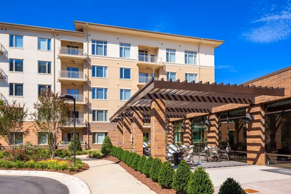 Exterior view of a multi-story senior living facility with beige and white walls, balconies, and large windows. In the foreground, there is a covered patio area with outdoor seating and a pergola structure. The area is landscaped with bushes, small trees, and a curved walkway under a clear blue sky.