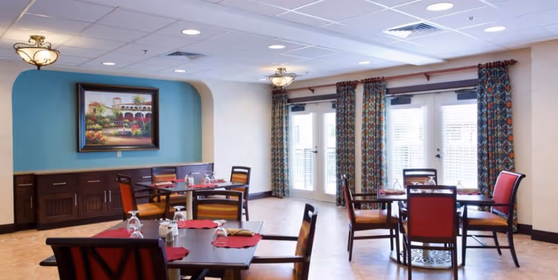 A dining room with several tables and chairs arranged neatly. Each table has red placemats and upside-down glasses. The room features large windows with patterned curtains, a blue accent wall with a framed painting, and ceiling lights providing bright illumination.