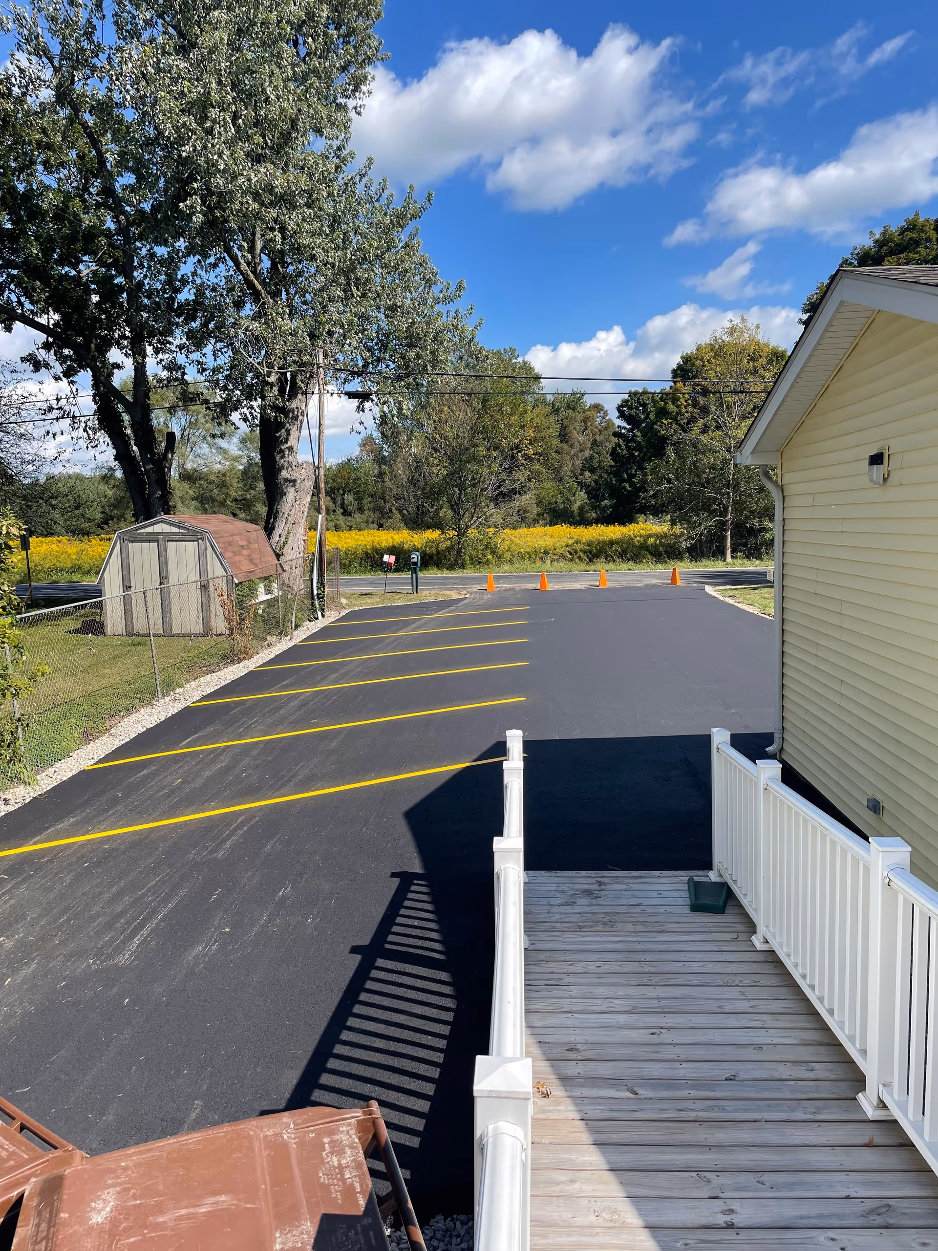 Freshly paved parking lot with yellow-striped spaces and orange cones beside a pale yellow building and wooden ramp, with a shed, trees and a field under a blue sky.