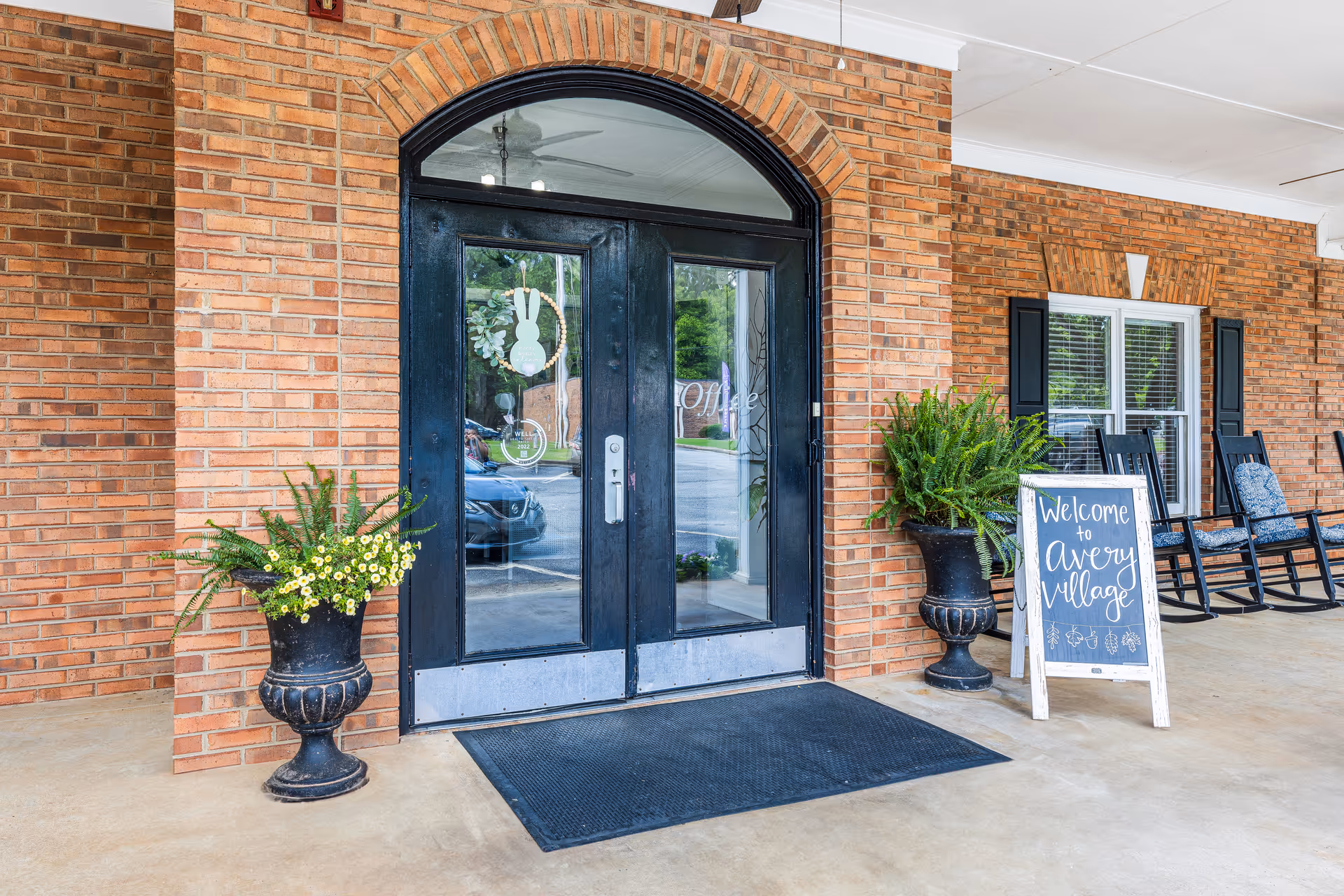 Entrance to Avery Village Assisted Living and Memory Care featuring double black glass doors with a wreath decoration, flanked by two large black planters with green plants and yellow flowers. A black mat is placed in front of the doors. To the right, there is a chalkboard sign that reads 'Welcome to Avery Village' and two black rocking chairs with blue cushions under a covered porch with brick walls.