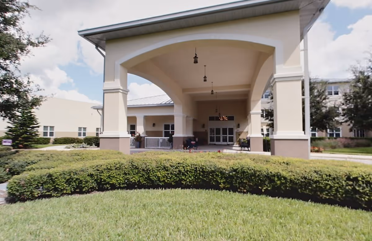 Front entrance of Bartram Lakes Assisted Living Facility - Brooks Rehabilitation showing a covered drop-off area with large columns, hanging lantern-style lights, and a well-maintained lawn and shrubbery in the foreground under a partly cloudy sky.