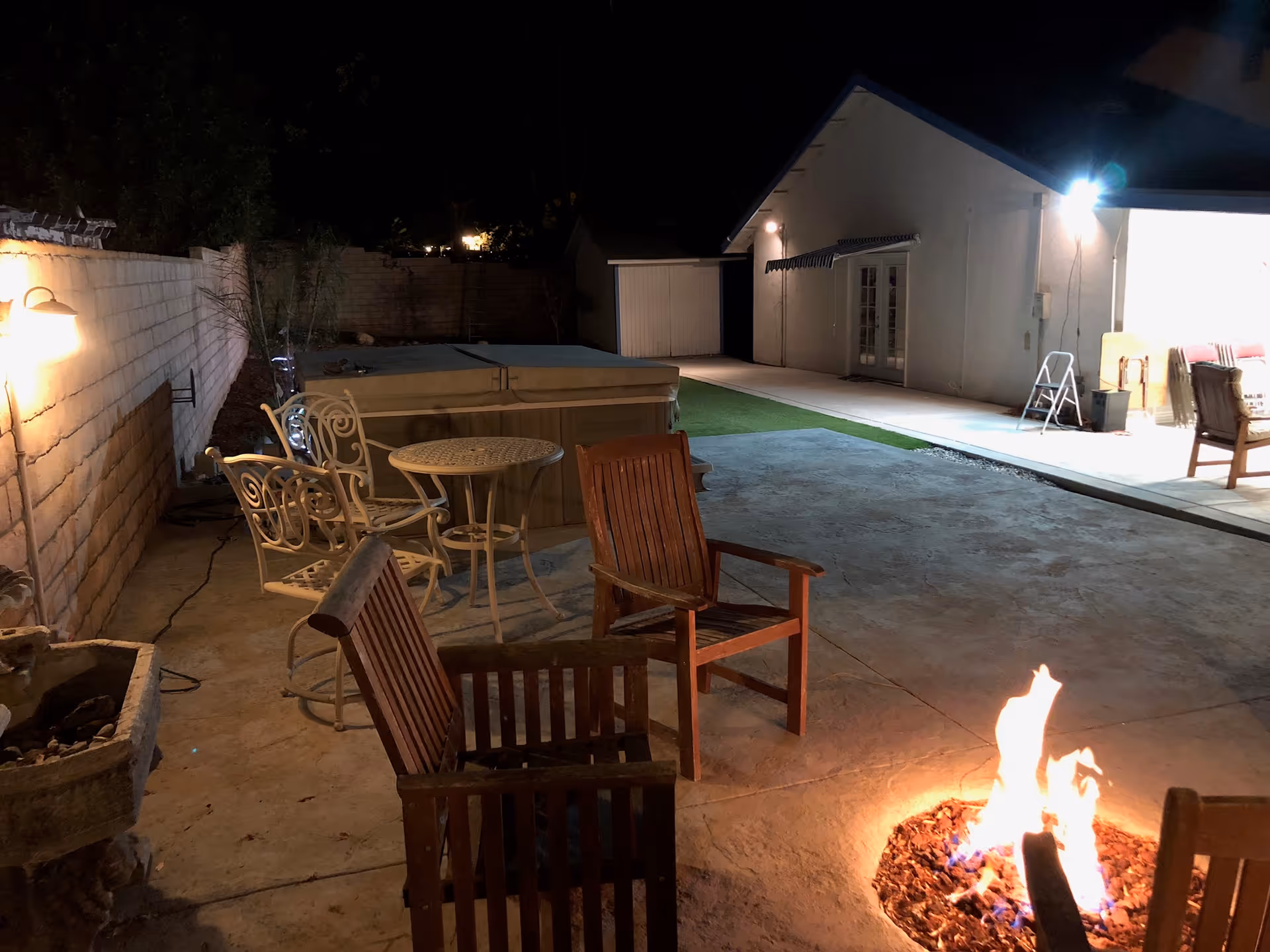 Outdoor patio area at night with a fire pit surrounded by wooden chairs, a small round metal table with matching chairs, and a hot tub against a brick wall. The area is lit by wall-mounted lights and a bright light near a building with a sloped roof and French doors.