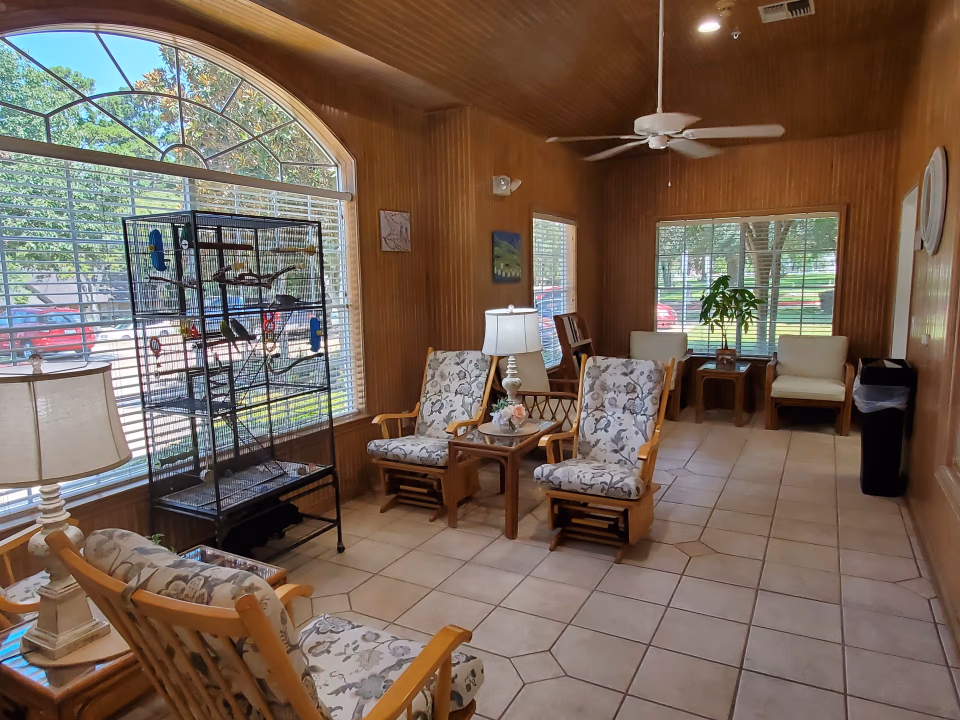 A cozy sitting area inside a senior living facility with wooden walls and tiled floor. The room features several cushioned wooden chairs arranged around small tables, a large birdcage with birds near a window, a ceiling fan, and large windows letting in natural light with views of trees and parked cars outside.