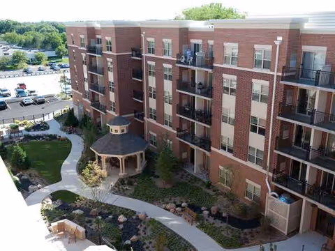 Exterior view of a brick senior living building with balconies, winding walkways, and a central gazebo.