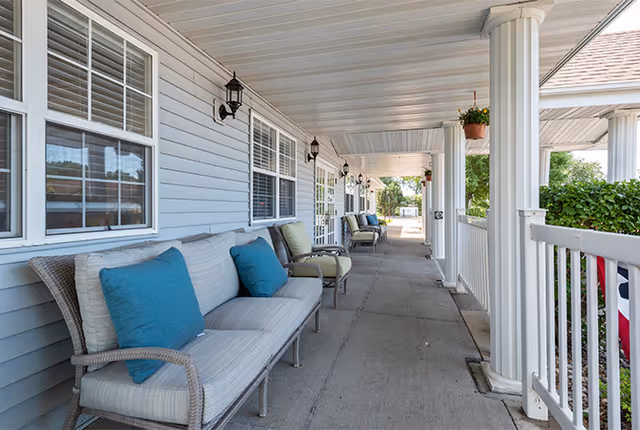 Covered outdoor porch area with cushioned wicker chairs and armchairs lined up against the building wall. The porch has white columns, hanging plants, and black wall-mounted lantern lights. There is a white railing on the right side and greenery beyond it.