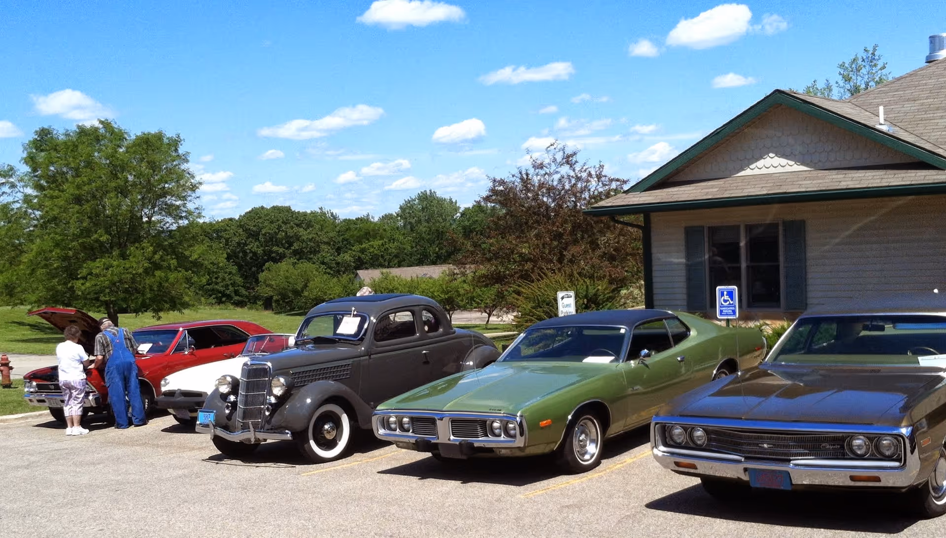 Several classic cars parked in front of a single-story building with a few people standing nearby on a sunny day.