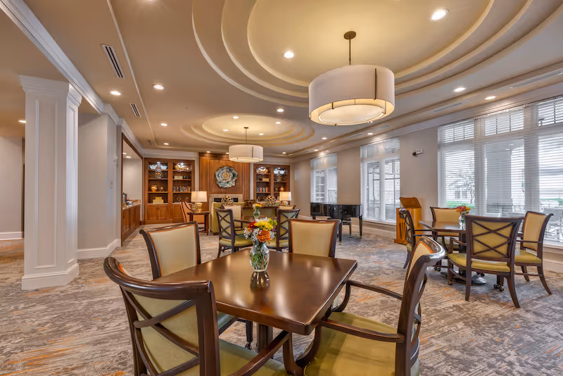 A spacious and elegant dining room with several wooden tables and chairs arranged neatly. Each table has a small vase with colorful flowers. The room features large windows with blinds allowing natural light to fill the space. The ceiling has decorative circular molding with modern pendant lights hanging down. In the background, there are built-in wooden cabinets and a grand piano near the windows.