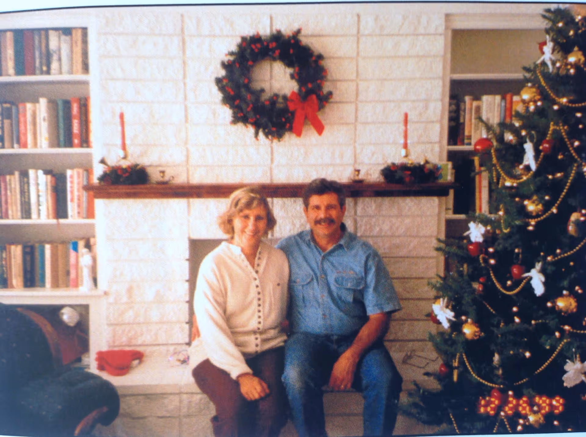 A middle-aged couple sitting together in a cozy living room decorated for Christmas. Behind them is a white brick fireplace with a wreath adorned with red berries and a red bow hanging above it. The mantel has two red candles and small festive decorations. To the right is a decorated Christmas tree with gold garlands, red ornaments, and white angel figurines. To the left is a bookshelf filled with books.