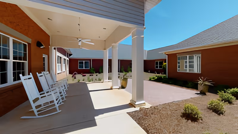 Covered porch with white rocking chairs overlooking a courtyard between red cottage buildings under a blue sky.