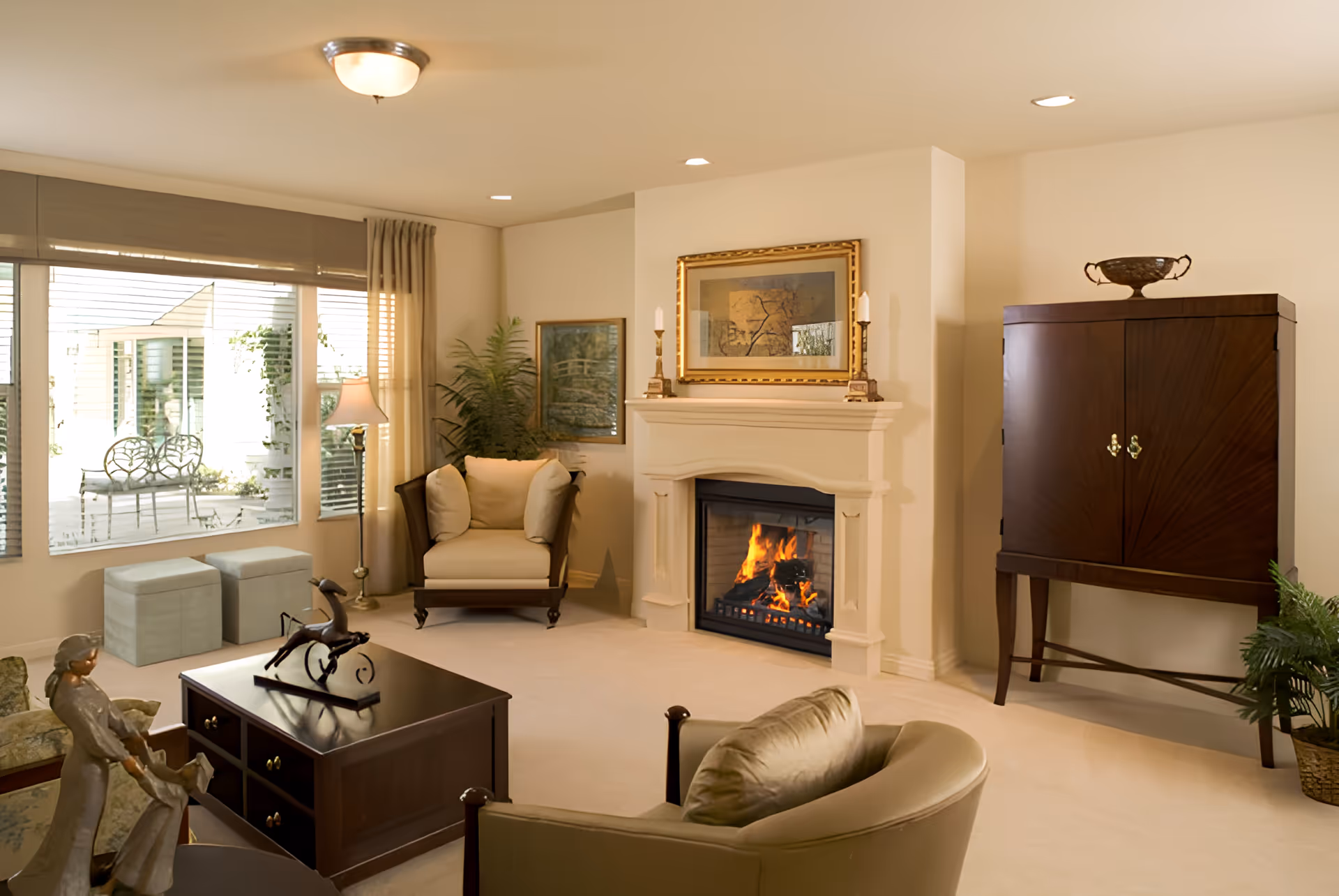 A cozy living room with a lit fireplace, upholstered chairs, a coffee table, large window, and a wooden cabinet.