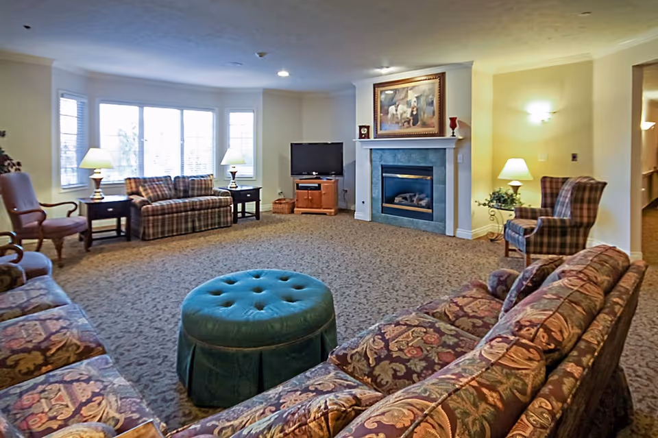 Spacious furnished living room with patterned sofas and armchairs, a central green ottoman, fireplace and TV.