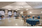 A spacious dining room with multiple round tables covered with teal tablecloths, each set with napkins and silverware. The room features chandeliers hanging from a white ceiling, large windows with curtains, and a wooden cabinet against the far wall.