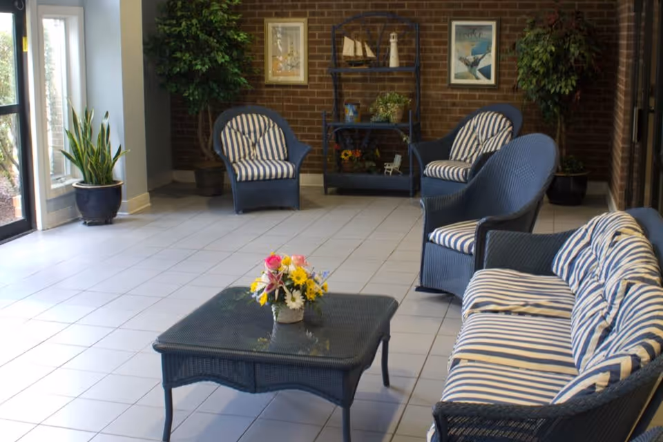 A cozy indoor seating area with blue wicker furniture including a sofa and three armchairs with blue and white striped cushions. A glass-top coffee table with a colorful flower arrangement is in the center. The room has a tiled floor, brick walls with framed pictures, potted plants, and a decorative shelf with model boats and plants.