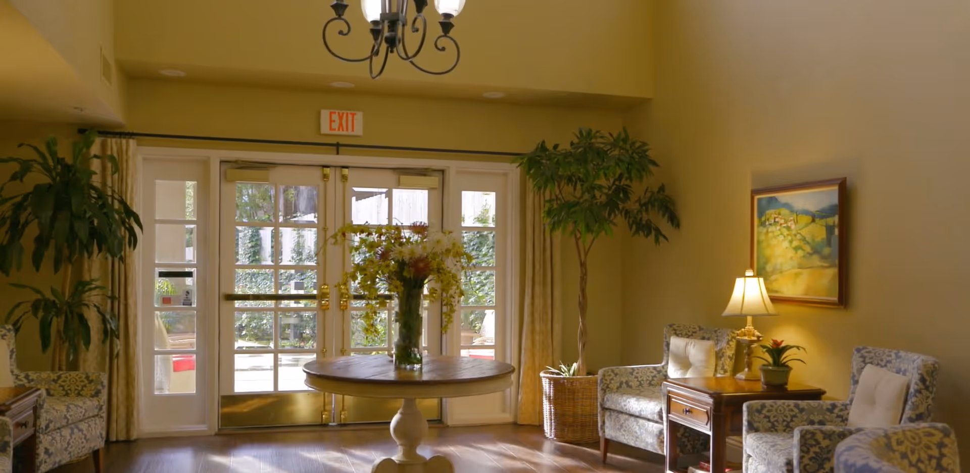 A cozy interior common area with two patterned armchairs, a wooden side table with a lamp and a small plant, a round wooden table with a vase of flowers in the center, large glass double doors with an exit sign above, and tall potted plants on either side.