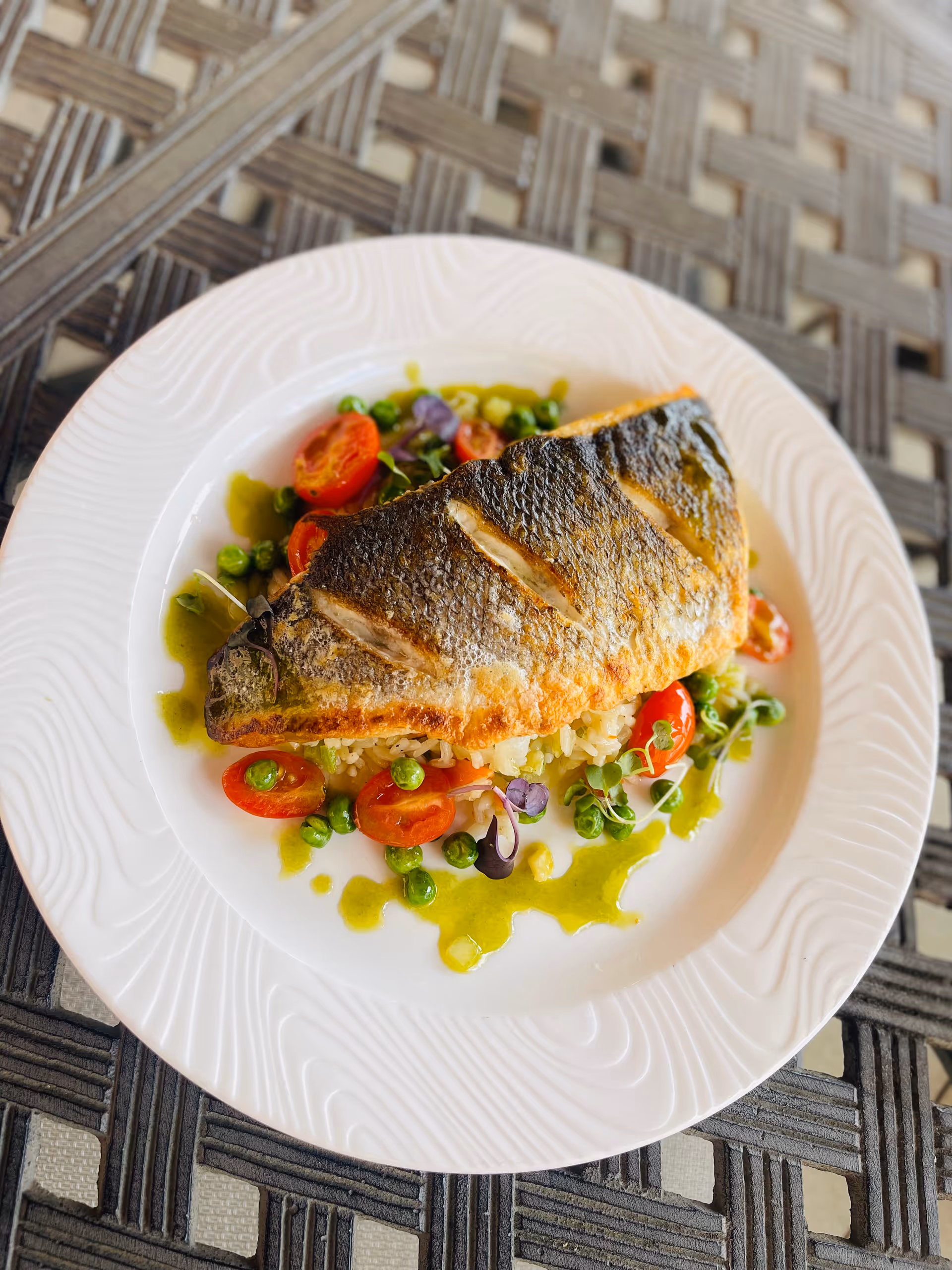 A white plate with a cooked fillet of fish served on a bed of rice, cherry tomatoes, green peas, and microgreens, drizzled with a green sauce. The plate is placed on a woven textured surface.