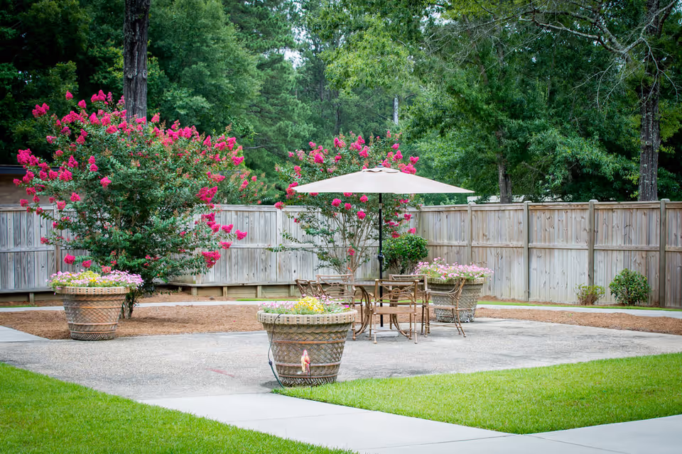 Outdoor patio area with a round table and several chairs under a large beige umbrella, surrounded by large flower pots with blooming flowers and vibrant pink flowering bushes, enclosed by a wooden fence and lush green trees in the background.