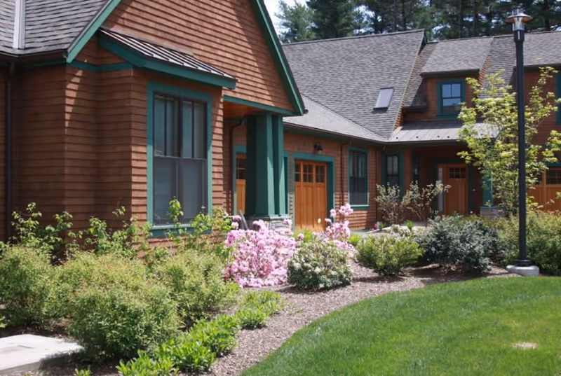 Exterior view of a building with brown wooden siding and green trim, surrounded by landscaped bushes and pink flowering plants, with a well-maintained lawn and a lamppost.