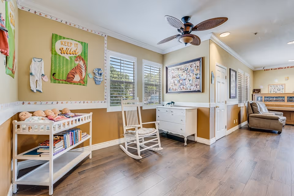 A bright and cozy interior room with wooden flooring, beige walls, and white trim. The room features a white rocking chair, a white dresser, and a shelf holding several baby dolls and books. On the wall above the dolls, there is a green poster with a tiger and the text 'KEEP IT WILD,' along with baby clothes hanging on the wall. The room has large windows with white shutters, a ceiling fan with wooden blades, and a comfortable brown armchair near the back. The space appears to be a common area or activity room in a senior living facility.