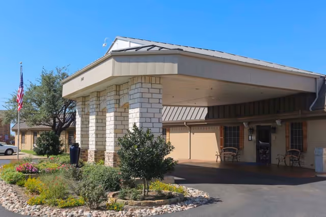 Covered entrance and driveway of a brick health and rehabilitation center with stone columns, landscaped planters, an American flag, and outdoor seating.