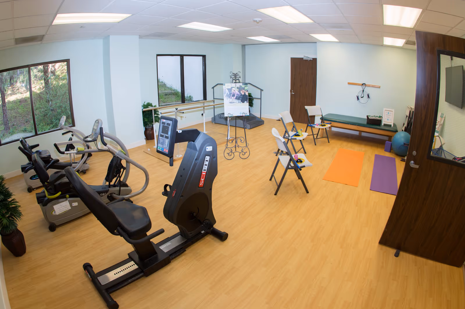 A bright exercise room with light wood flooring and pale blue walls. The room contains several pieces of exercise equipment including stationary bikes and a recumbent bike. There are three folding chairs with small weights on them, two yoga mats on the floor, a bench, and a large exercise ball. Two large windows show greenery outside. A door is open on the right side of the image.