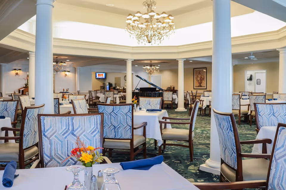 A spacious dining room with multiple tables covered in white tablecloths, each set with blue napkins, glassware, and small floral centerpieces. The room features elegant columns, chandeliers, and a grand piano in the background. The chairs have patterned upholstery and wooden frames, and the carpet is green with a decorative pattern.