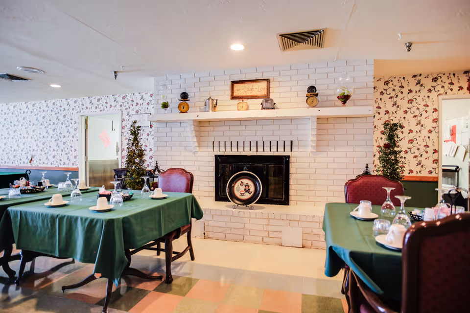 A dining room with tables covered in green tablecloths set with cups, glasses, and napkins. The room features a white brick fireplace decorated with small vintage scales and a framed sign above it. The walls have floral wallpaper, and there are red cushioned chairs around the tables.