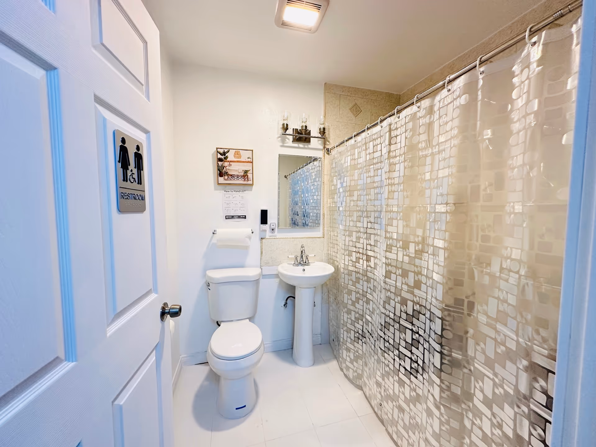Small bathroom with toilet, pedestal sink, and patterned shower curtain in a senior facility.