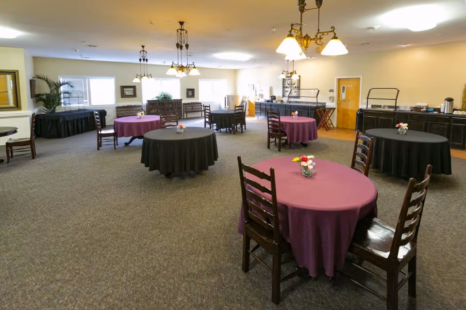 A spacious dining room with round tables covered in purple and black tablecloths, each adorned with small floral centerpieces. Wooden chairs surround the tables. The room has beige walls, carpeted floors, multiple windows letting in natural light, and several hanging light fixtures. There is a counter area along one wall with coffee and beverage supplies.