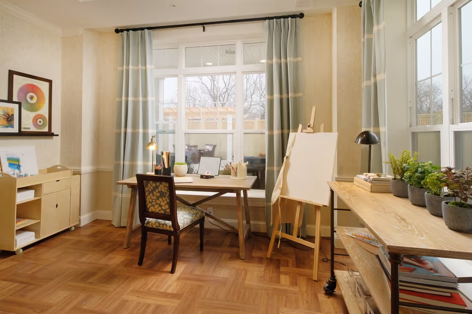 A bright and cozy art room with a wooden desk and chair in front of large windows with light blue curtains. The desk has art supplies including pencils, brushes, and a small plant. To the right, there is an easel with a blank canvas and a wooden table holding several potted plants and books. The room has light-colored walls and a wooden parquet floor.