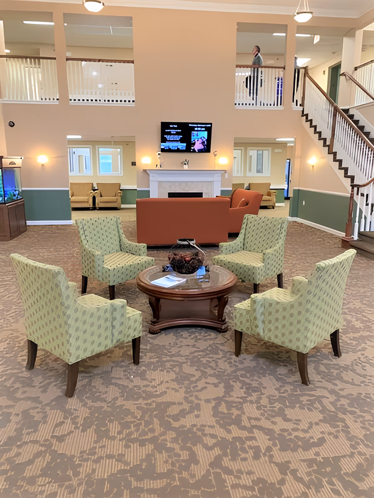 A spacious living area in a senior living facility with five patterned green armchairs arranged around a round wooden coffee table. Behind the chairs is a red sofa facing a white fireplace with a mounted flat-screen TV above it. The room has beige walls with green wainscoting, carpeted floors, and a staircase on the right side leading to an upper level with white railings. There is also a fish tank on the left side and additional seating visible through two open doorways.