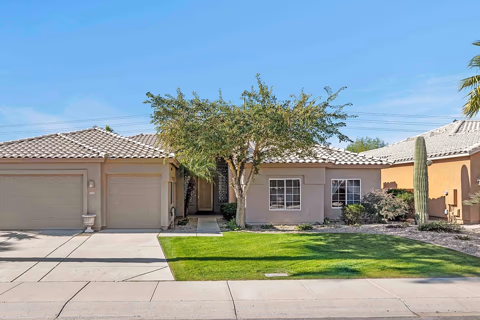 Single-story stucco house with a two-car garage, tiled roof, central entryway framed by a tree, a green front lawn and desert landscaping including a tall cactus.