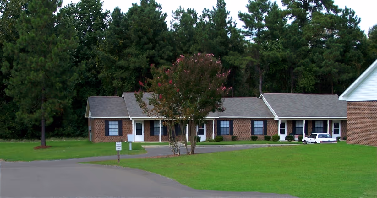 Exterior view of a single-story brick building with multiple doors and windows, surrounded by green grass and trees in the background. A small tree with pink flowers is in front of the building, and a white car is parked near one of the doors.