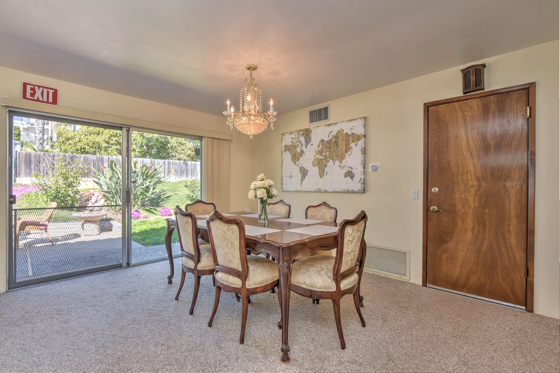 A dining room with a wooden table and six upholstered chairs. A vase with white flowers is placed on the table. A chandelier hangs from the ceiling. There is a large world map artwork on the wall and a wooden door to the right. Sliding glass doors on the left open to a garden with greenery and flowers. An exit sign is visible above the sliding doors.