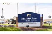Outdoor entrance sign reading 'ManorCare Health Services Nursing & Rehab' on a landscaped lawn with the facility and vehicles visible behind it.