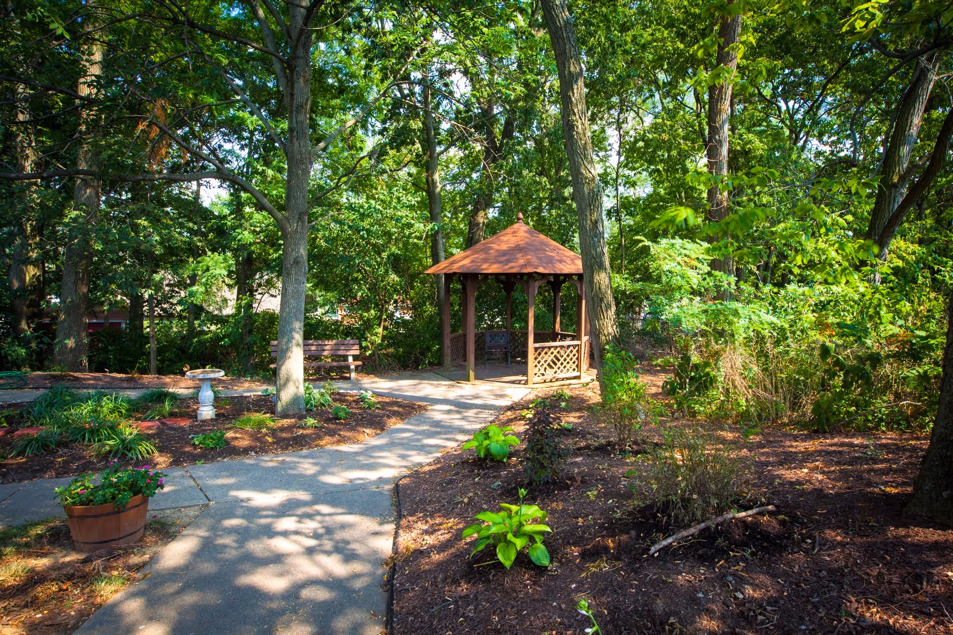A peaceful garden area with a paved walkway leading to a wooden gazebo surrounded by trees and plants. There is a bench and a birdbath along the path, with sunlight filtering through the foliage.