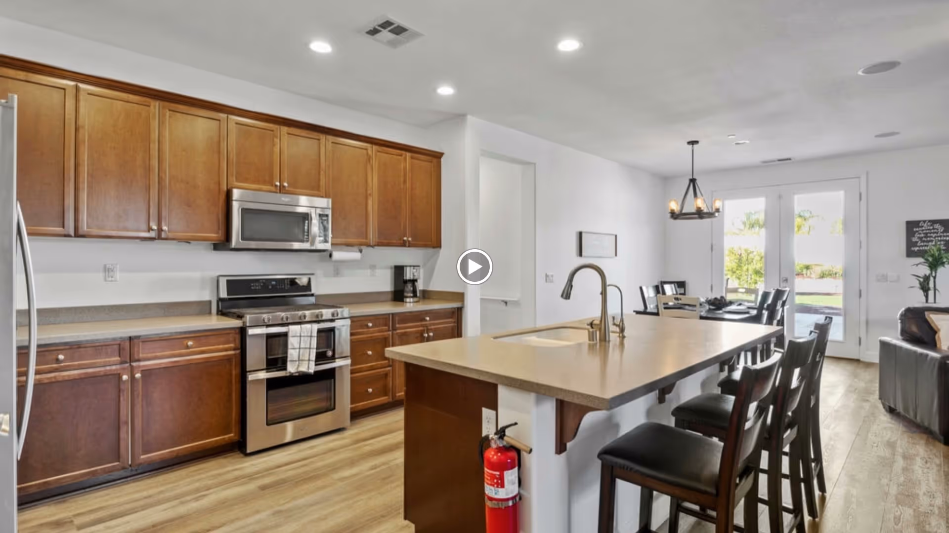 Modern kitchen with wooden cabinets, stainless steel appliances including a refrigerator, stove, and microwave. A large kitchen island with a sink and faucet has several dark wooden bar stools. The kitchen opens into a dining area with a table and chairs near glass double doors leading outside. The floor is light wood, and the walls are white with recessed lighting in the ceiling.