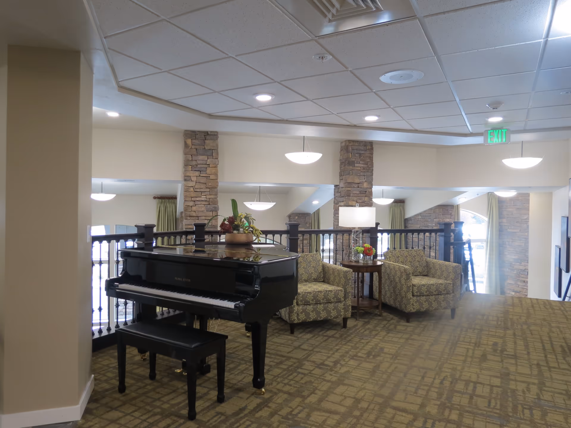 Interior common area with a black grand piano and matching bench, two patterned armchairs, a small round table with a lamp and flower arrangement, stone pillars, hanging ceiling lights, and large windows with green curtains.
