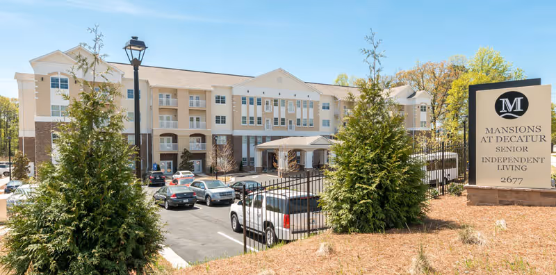 Exterior view of a multi-story senior living facility building with a parking lot in front. There are several cars parked, trees, and a large sign that reads 'Mansions at Decatur Senior Independent Living 2677'. The sky is clear and the area is well-maintained.