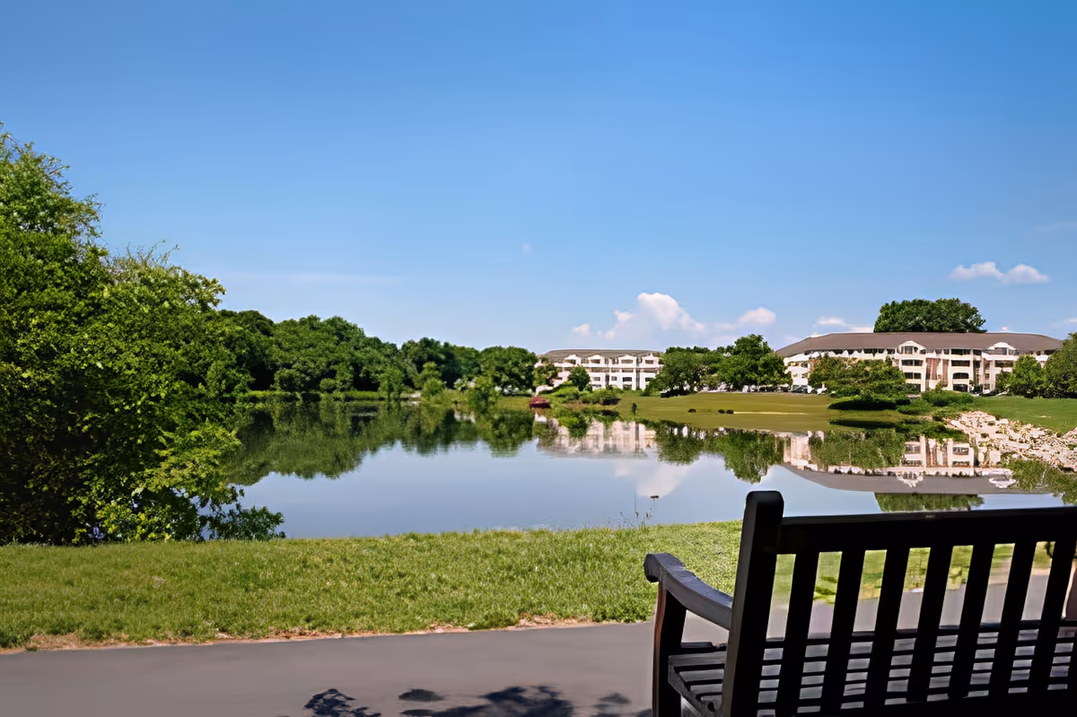 A peaceful outdoor scene featuring a calm lake with clear reflections of trees and a large building in the background. In the foreground, there is a wooden bench facing the lake, situated on a paved path with green grass surrounding it under a clear blue sky.