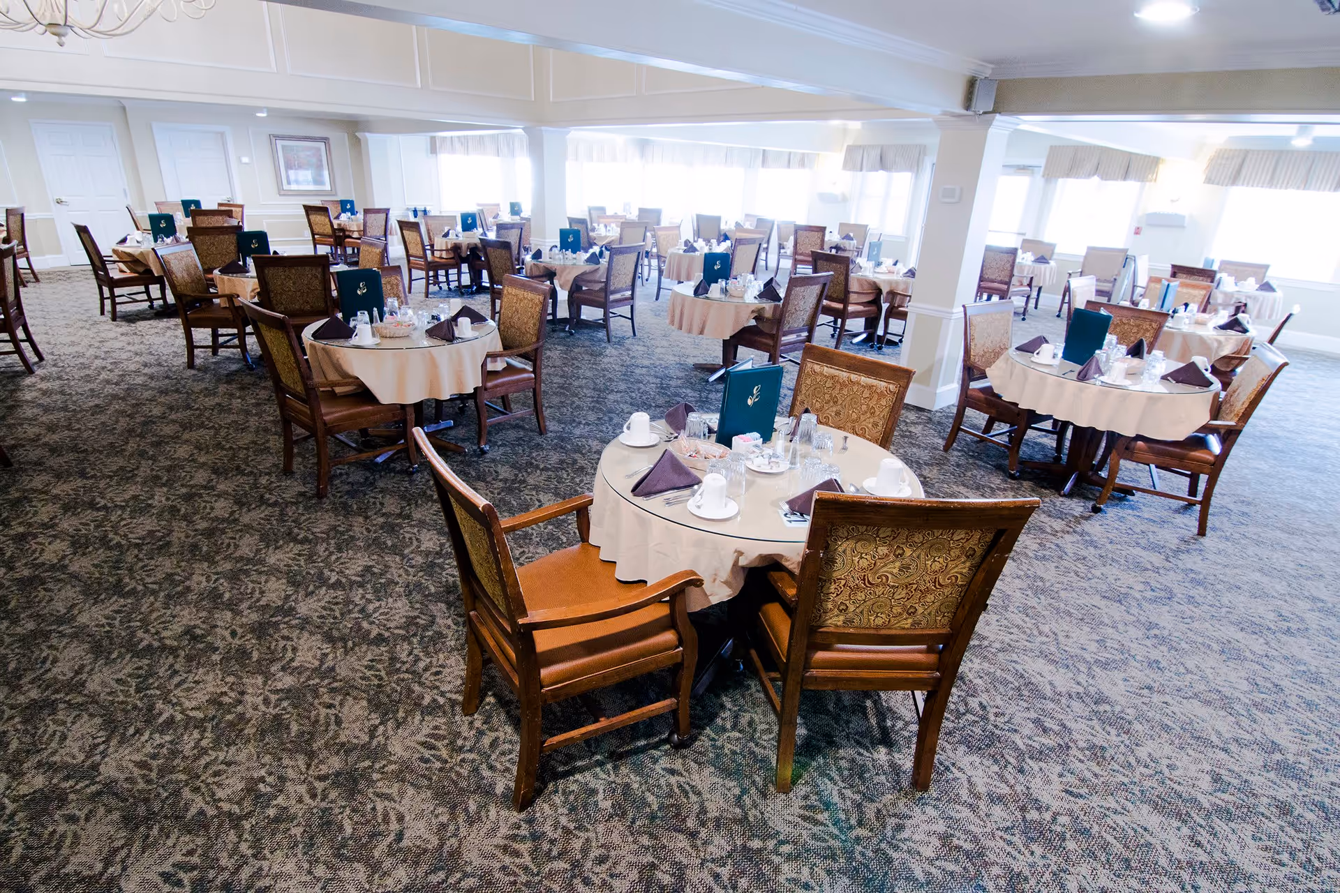 A spacious dining room with multiple round tables covered with beige tablecloths, each set with cups, glasses, napkins, and menus. The room has patterned carpet flooring, large windows allowing natural light, and upholstered wooden chairs around each table.