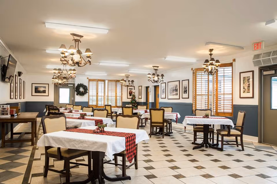 Bright dining room with multiple tables and chairs, chandeliers, checkered tile floor and holiday decorations.