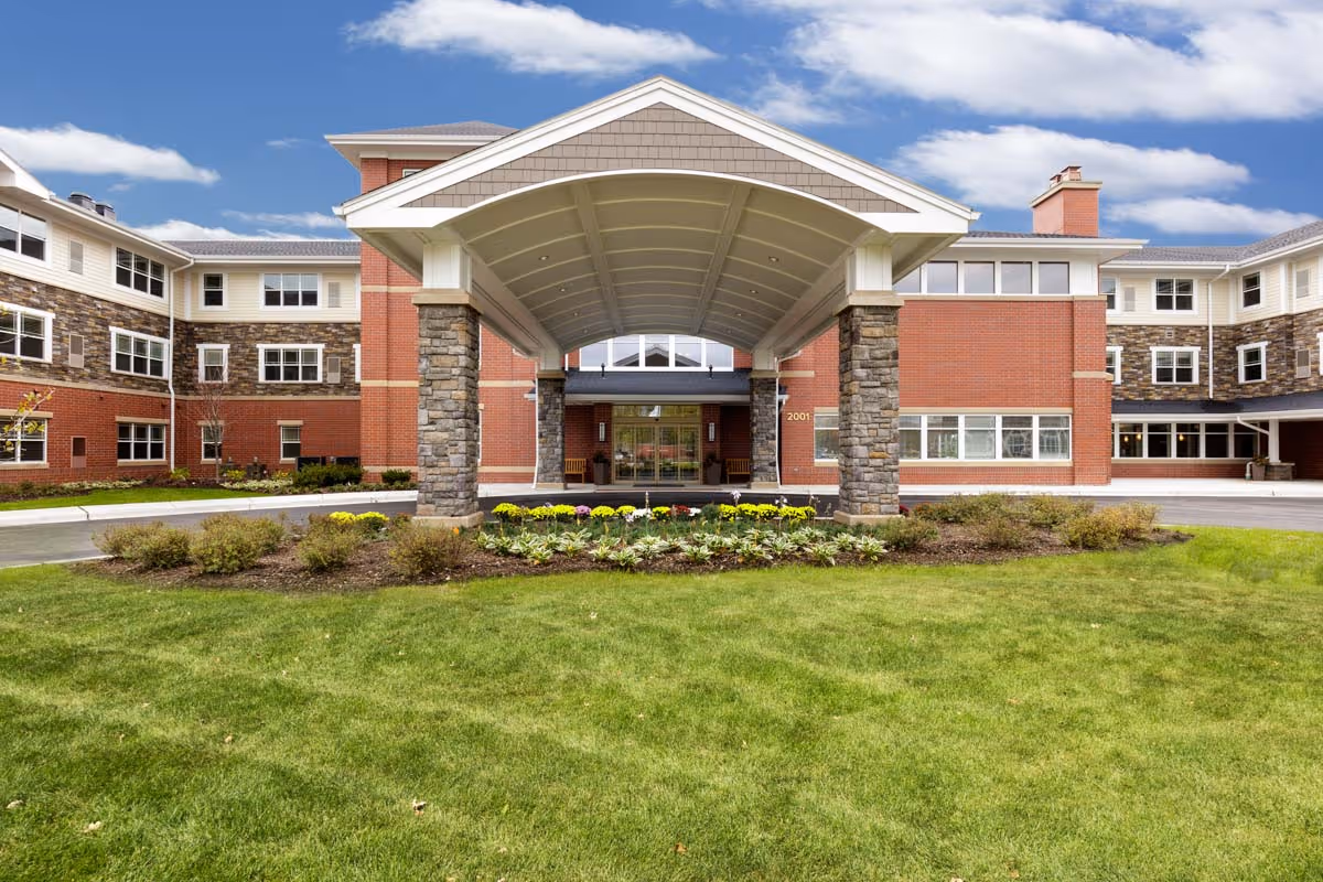 Front exterior view of Clarendale of Algonquin, a multi-story senior living facility with a covered entrance supported by stone pillars, surrounded by landscaped greenery and a clear blue sky with some clouds.