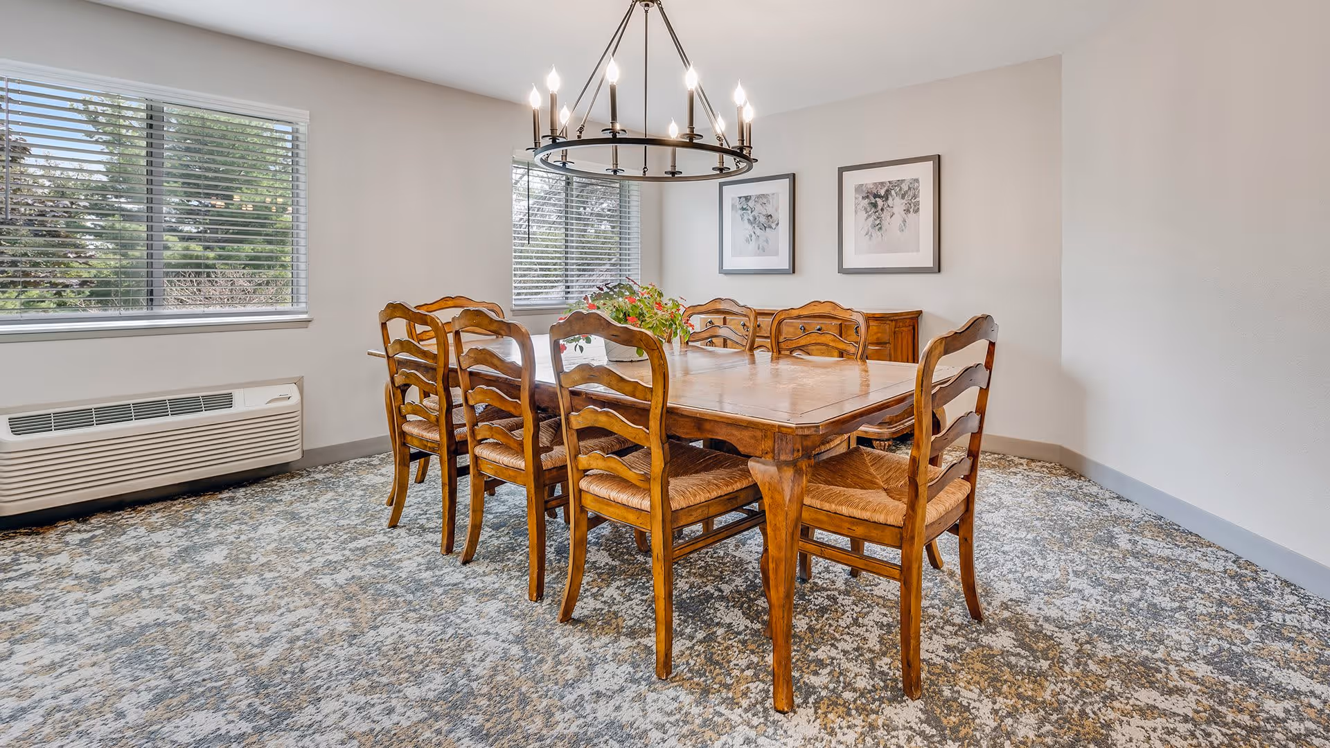A dining room with a wooden table and eight matching wooden chairs with woven seats. The room has two large windows with blinds, a patterned carpet, a chandelier hanging above the table, and two framed floral artworks on the wall.
