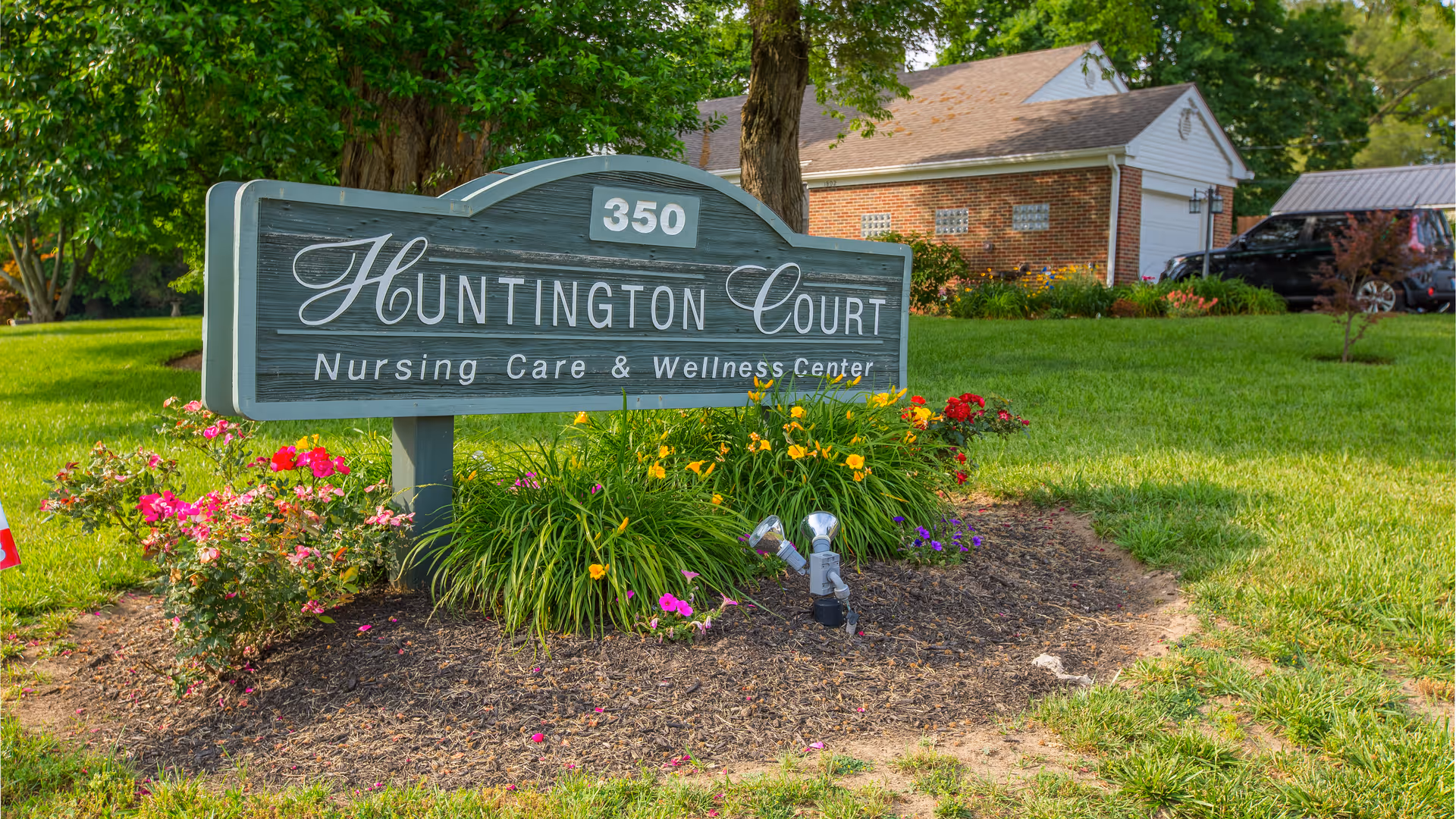 A wooden sign for Huntington Court Nursing Care & Wellness Center with the number 350 on top, surrounded by colorful flowers and green grass, with a brick building and trees in the background.