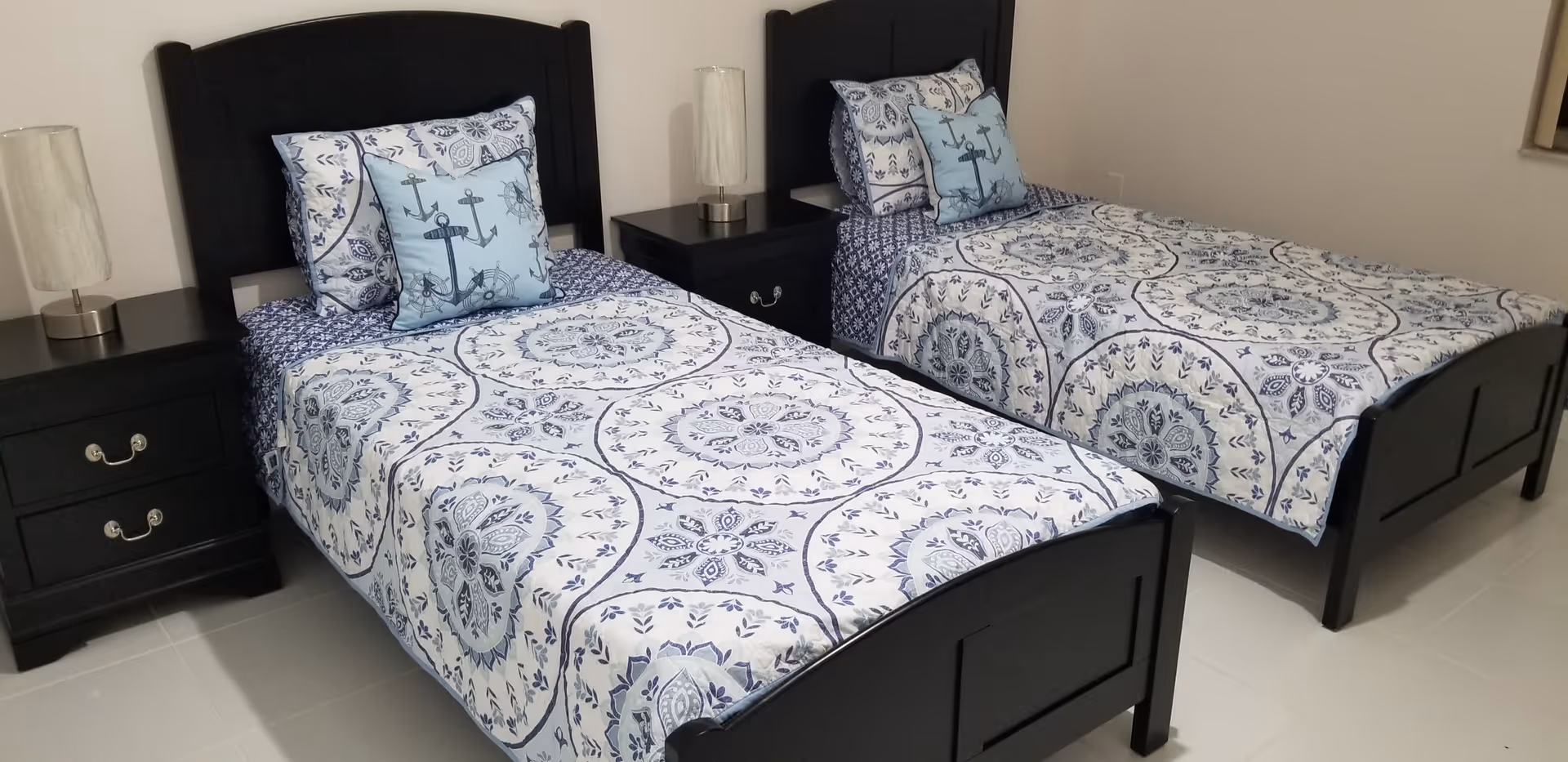 A bedroom with two single beds featuring black wooden headboards and footboards. Each bed is made with matching blue and white patterned bedspreads and pillows, including a decorative pillow with an anchor design. Between the beds are two black nightstands, each with a lamp on top. The room has light-colored tiled flooring and neutral-colored walls.